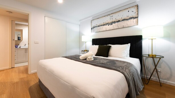 Een netjes opgemaakt bed met hoofdbord, twee bedlampen, een aan de muur hangend kunstwerk en een zichtbare badkamedeur.