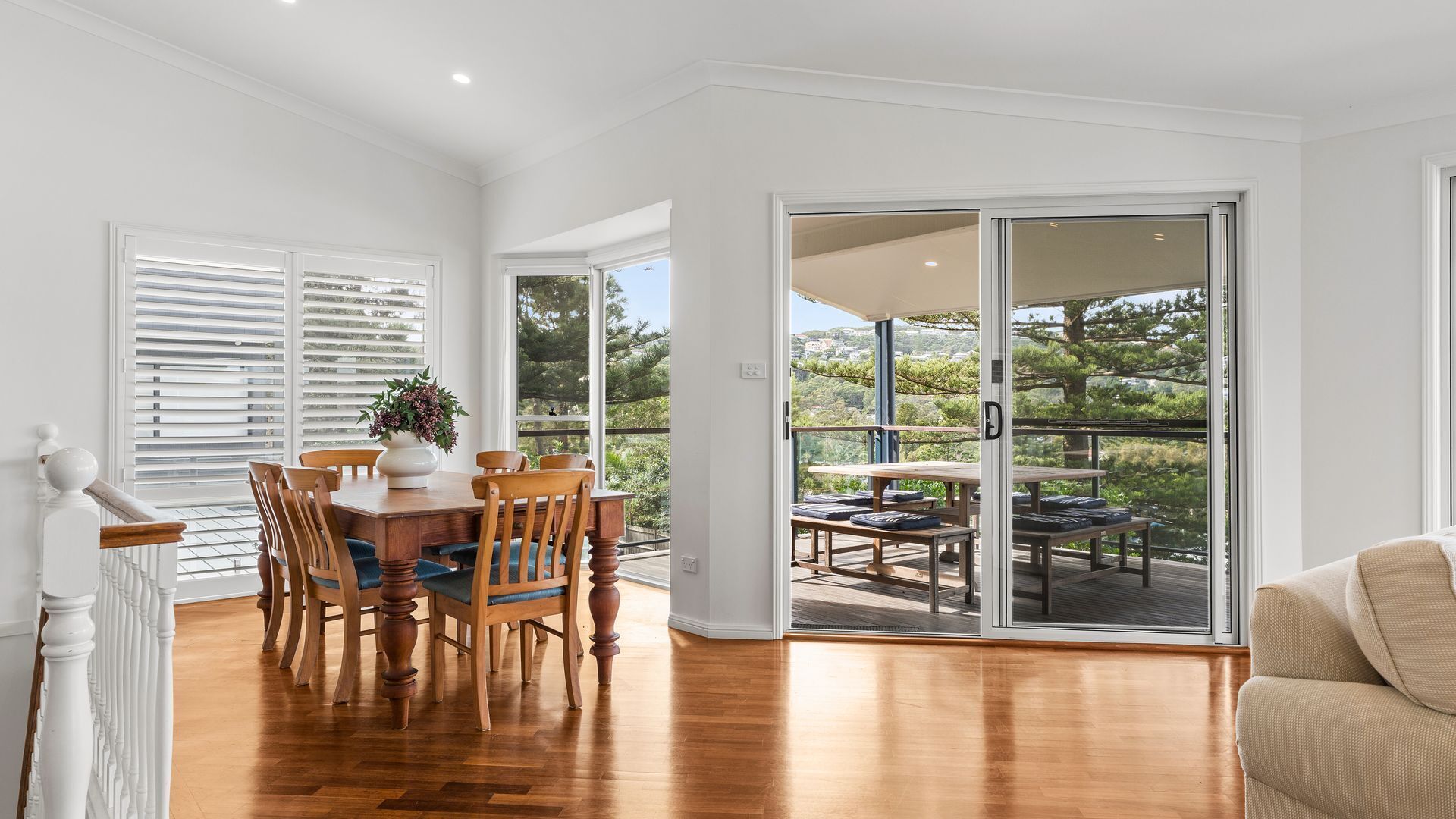 A dining area with a wooden table and chairs, a vase with flowers, and a view of a balcony with outdoor furniture.