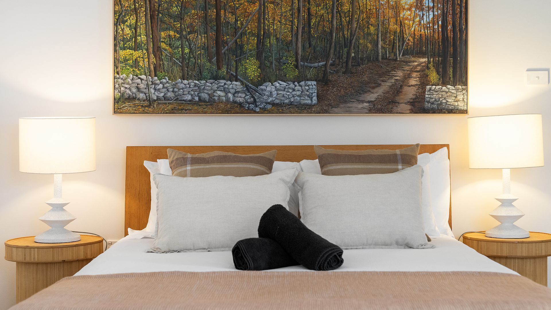 A bed with white bedding, two pillows, and a black towel, flanked by two bedside tables with lamps, in a room with a painting of a forest path above the bed.