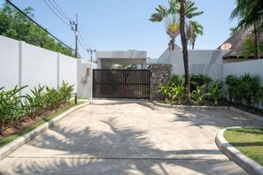 A gated driveway leading to a residential property with a white wall, a black gate, and a palm tree. - Anchan Villas Phuket - Private 4 BR Signature Pool Villa by Escape Villas (Phuket)