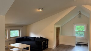 Una habitación con un sofá negro, una mesa blanca, un techo inclinado y una ventana con vista a árboles.