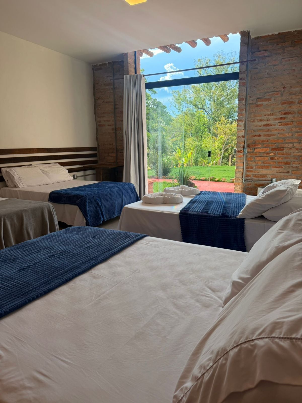 A hotel room with two beds, a large window with a view of greenery, and a brick wall.