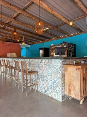 A bar area with wooden chairs, a counter with a beverage dispenser, and hanging pendant lights.