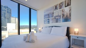 Habitación de hotel con una cama grande, una mesita de noche, una pared decorada con imágenes de la ciudad y vistas a edificios a través de amplias ventanas.