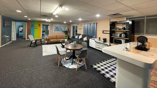 En moderne stue med sofa, spisebord og stole, et køkkenområde med hårde hvidevarer, og en trappe, der fører til et andet niveau.