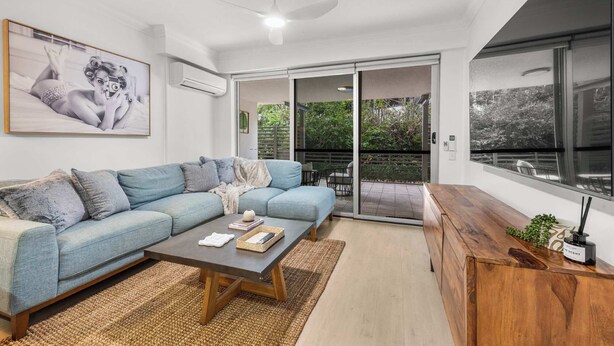 A modern living room with a sofa, coffee table, and a wooden cabinet. There is a sliding glass door leading to an outdoor area with a view of greenery. - Spacious 3BR Retreat | Pool, Gym & Parking (Bowen Hills)