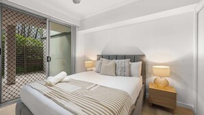 A bedroom with a bed, two bedside lamps, a nightstand, and a sliding glass door leading to a balcony. - Spacious 3BR Retreat | Pool, Gym & Parking (Bowen Hills)