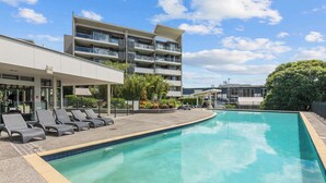A swimming pool with lounge chairs, a multi-story building with balconies, and a clear sky. - Spacious 3BR Retreat | Pool, Gym & Parking (Bowen Hills)