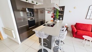 Une cuisine moderne dotée d’appareils électroménagers en acier inoxydable, un îlot central avec des tabourets de bar, et un canapé rouge dans le salon attenant.