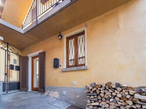 Un bâtiment à l’extérieur jaune, avec une porte en bois, une fenêtre ornée d’une grille métallique et une pile de bois de chauffage.