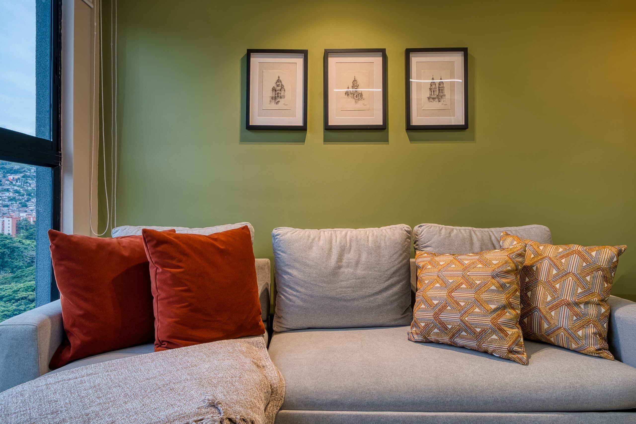 A room with a grey sofa, three framed pictures on the wall, and a window with a view of buildings.