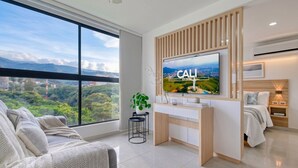 A modern hotel room with a large window, a flat-screen TV mounted on a wooden panel, a bedside table with a lamp, and a bed with a white comforter. - Edificio Miro Guadalupe (Cali)