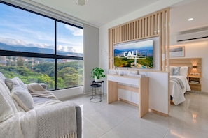 A modern hotel room with a large window, a flat-screen TV mounted on a wooden panel, a bedside table with a lamp, and a bed with a white comforter.