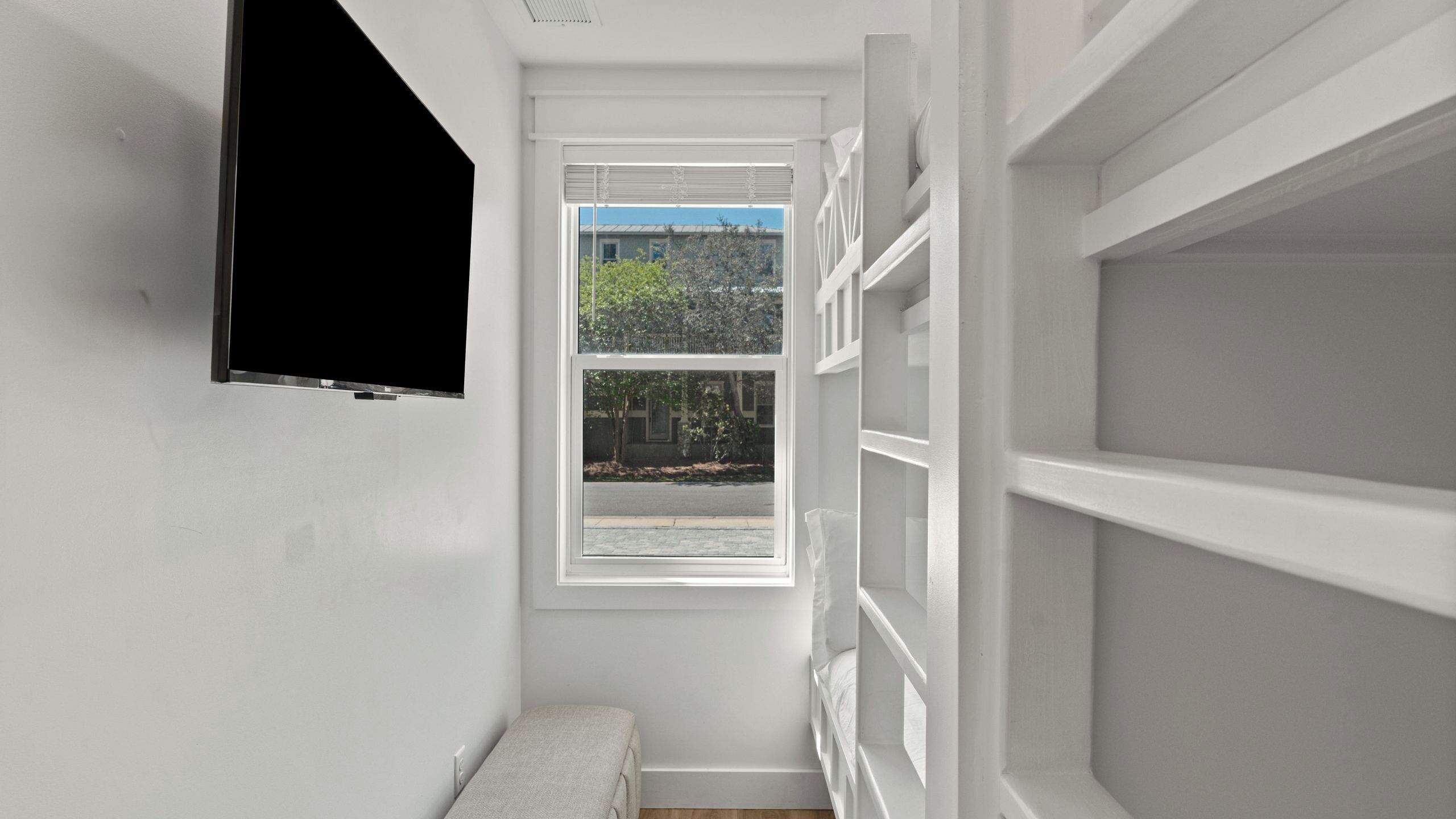 Ein schmaler Korridor mit weißen Wänden, einem Fenster mit Jalousien und einem angebrachten Fernseher.