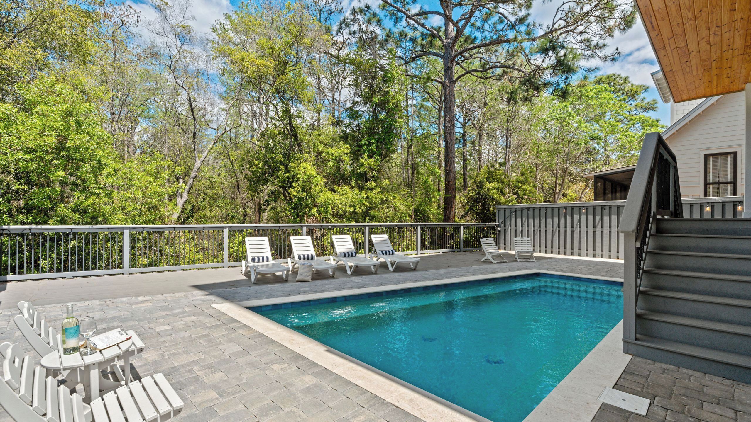 A swimming pool with lounge chairs, a table with a bottle and glass, and a wooden deck with a railing.