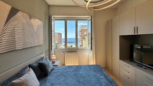 A bedroom with a bed, a TV, a window with a view of buildings and the sea, and a modern light fixture. - Double One Beach & City (Recco)