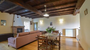 A spacious living area with a wooden ceiling, a fireplace, a sofa, a dining table with chairs, and a kitchen area in the background.