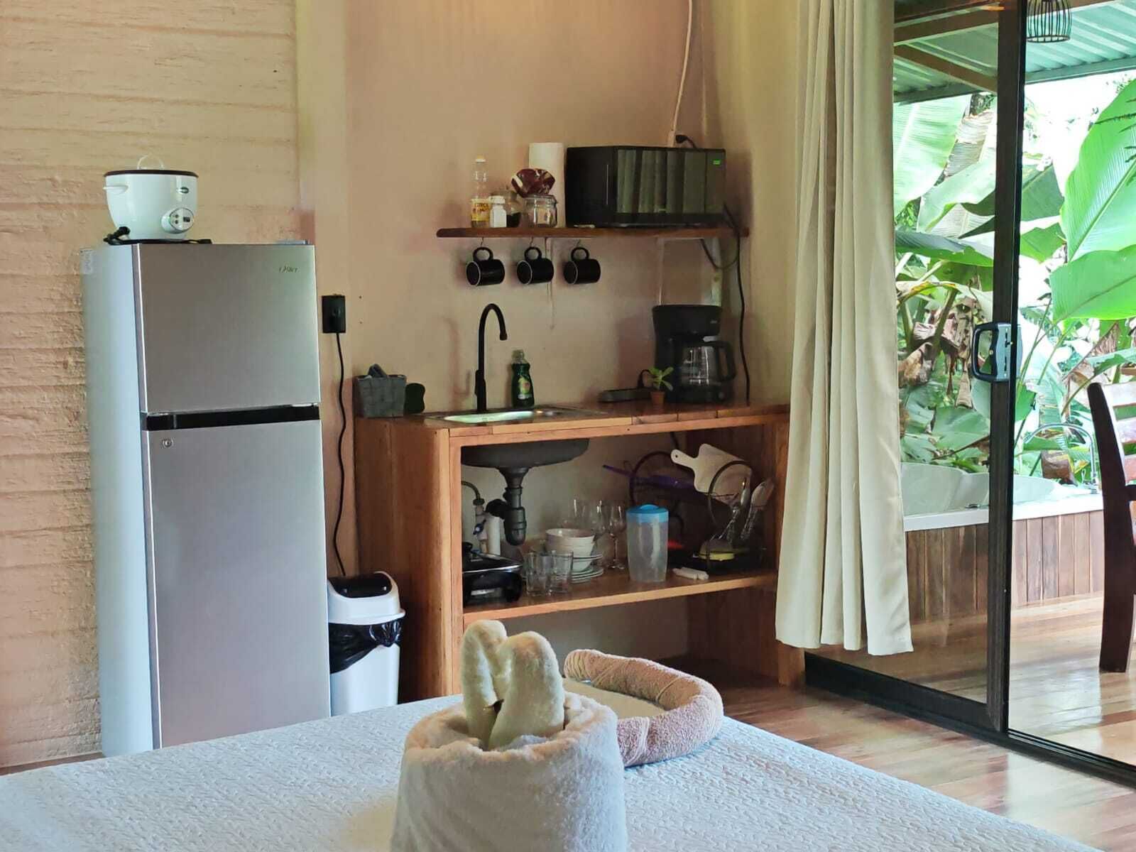 A compact kitchen with a refrigerator, coffee maker, and a sink. There is a small shelf with mugs and a window with a view of greenery.