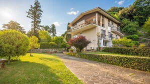 Ein zweistöckiges Gebäude mit Balkon, umgeben von Grünpflanzen und einem Kiesweg.