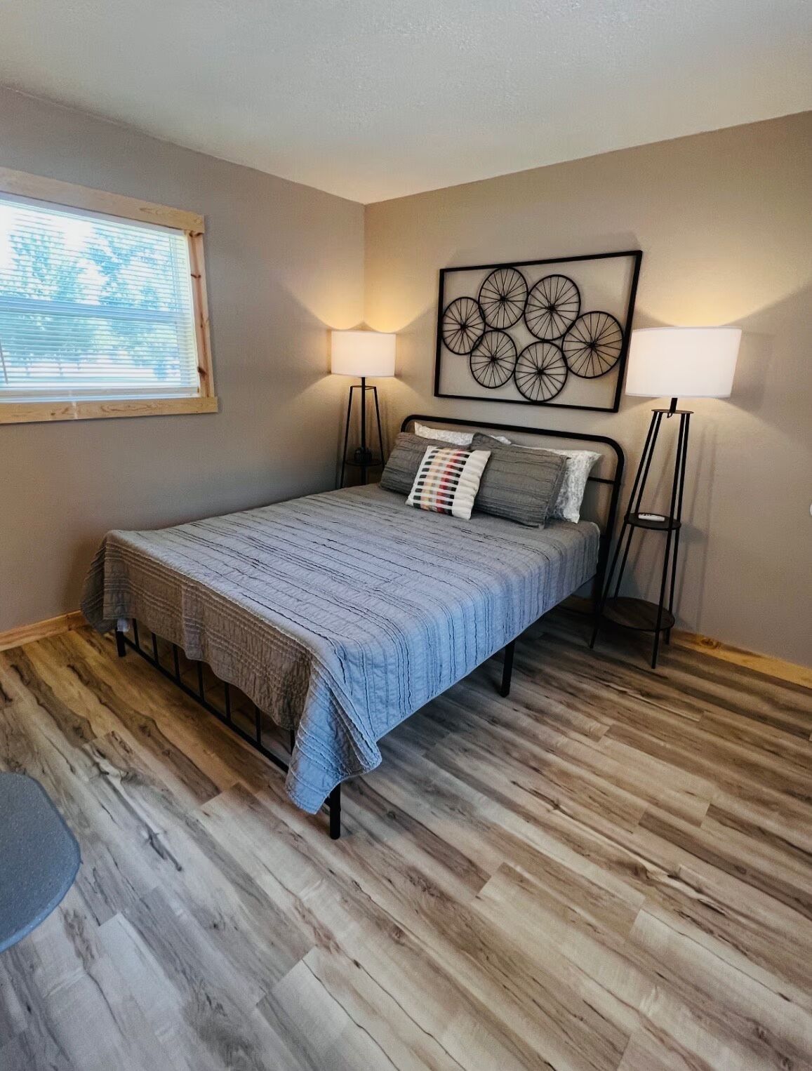 A bedroom with a bed, two bedside lamps, a window with blinds, a wall art piece, and a wooden floor.