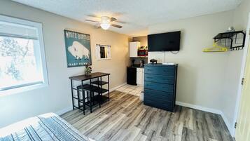 Une pièce avec un ventilateur de plafond, un téléviseur fixé au mur, une petite table avec des chaises, une commode et une affiche accrochée au mur.