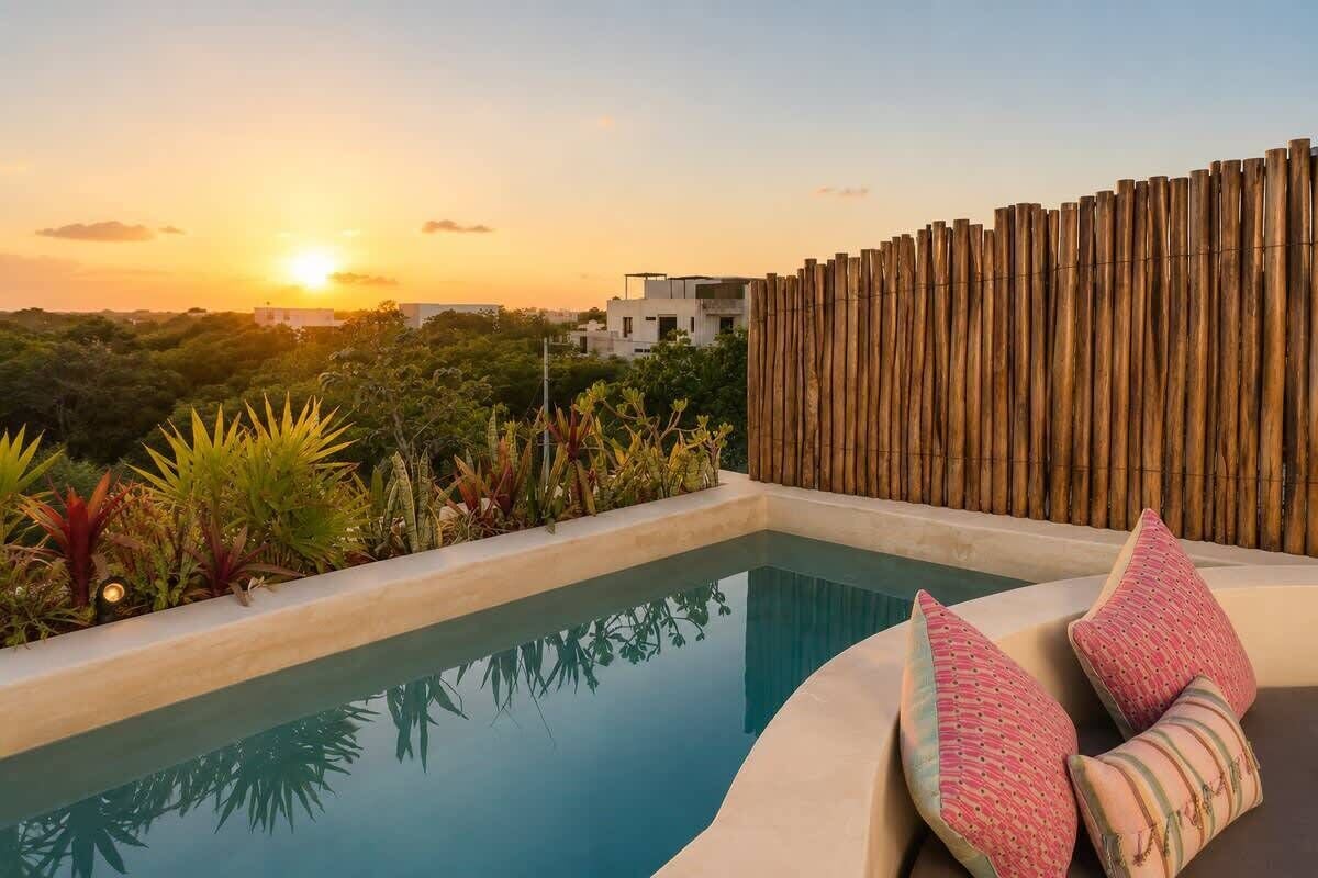 A poolside area with a wooden fence, a cushioned seating area, and a view of the sunset.