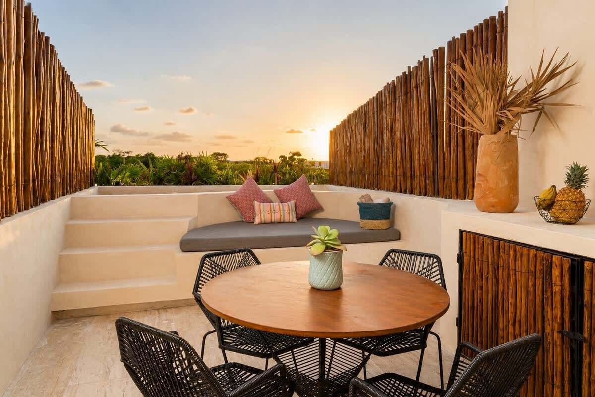 A terrace with a round wooden table, black wicker chairs, a cushioned bench with pillows, a bamboo wall, and a view of the sunset.