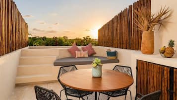A terrace with a round wooden table, black wicker chairs, a cushioned bench with pillows, a bamboo wall, and a view of the sunset.