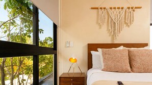 Een slaapkamer met een groot raam, een nachtkastje met een lamp, een macramé wanddecoratie en een bed met gekleurde kussens.