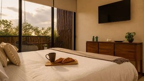 Un dormitori amb un llit, una taula de cafè amb croissants, una televisió i vistes a arbres a través de grans finestres.