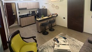 Una cucina moderna con mobili in legno, un'isola centrale con sgabelli da bar, una zona pranzo con divano e tavolino, e un corridoio che porta a una porta.