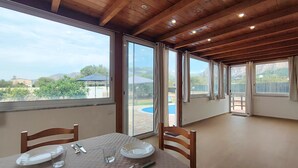 Ein Essbereich mit Tisch für zwei Personen, großen Fenstern und Blick auf einen Swimmingpool und Grünflächen.
