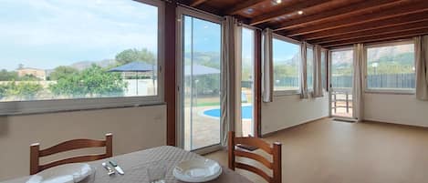 Comedor con mesa dispuesta para dos, ventanas amplias y vistas a una piscina y vegetación.