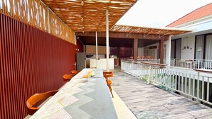 A covered outdoor dining area with a long table and chairs, a wooden wall, and a balcony with a railing.
