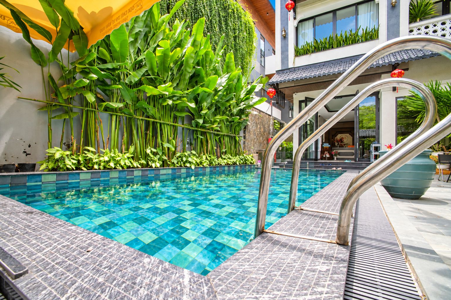 Una piscina con diseño de baldosas en damero, rodeada de exuberante vegetación y un edificio moderno con balcón.