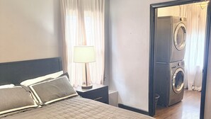 Une chambre à coucher comprenant un lit, un ventilateur de plafond, une table de chevet, une lampe et donnant sur une buanderie.