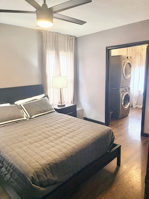 A bedroom with a bed, a ceiling fan, a nightstand, a lamp, and a view of a laundry area.
