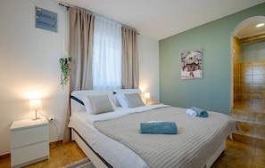 Un dormitorio con cama, mesitas de noche, un enchufe en la pared, una ventana con cortinas, una obra de arte en la pared y una zona de ducha.