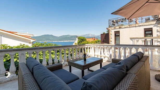 Un balcón con mobiliario de mimbre, una mesa de vidrio y vistas al mar y las montañas.