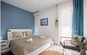 A bedroom with a bed, a chair, a picture on the wall, and an air conditioning unit.