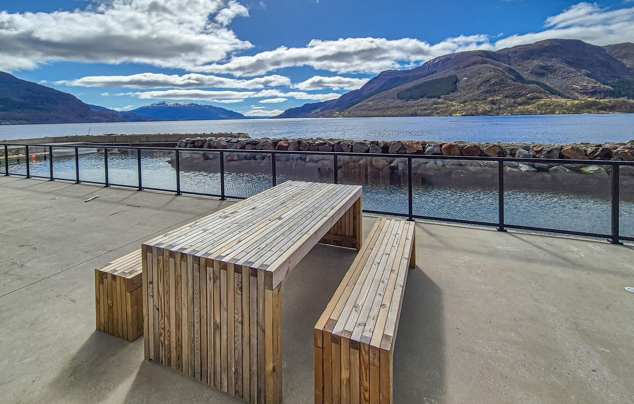 A wooden bench and table on a concrete deck with a railing overlooking a lake and mountains.