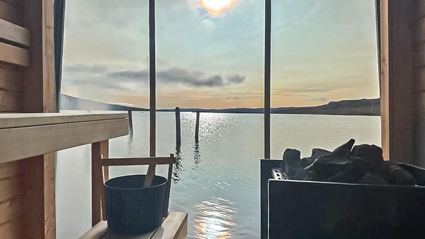 A room with a large window overlooking a body of water, a wooden bench, and a bucket. - Cozy home in Vøringsfoss with sauna (Vøringsfoss)