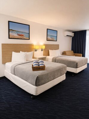 Ein Hotelzimmer mit zwei Betten, einem Holzkopfende, einem Nachttisch mit Lampen, einem kleinen Tisch und Blick auf den Strand.