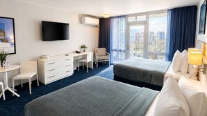 Ein Hotelzimmer mit zwei Betten, einem Schreibtisch, einem Stuhl, einem Fernseher und einem großen Fenster mit Vorhängen.