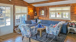 L’intérieur d’une cabane en rondins, avec un canapé, des fauteuils à motifs floraux, une table et une fenêtre donnant sur un paysage.
