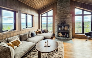 A cozy living room with a stone fireplace, large windows, and a wooden ceiling.