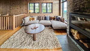Un salon chaleureux doté d’un plancher en bois, d’une table basse ronde, d’un canapé d’angle agrémenté de nombreux coussins, d’une cheminée en pierre et de grandes fenêtres offrant une vue sur la verdure.