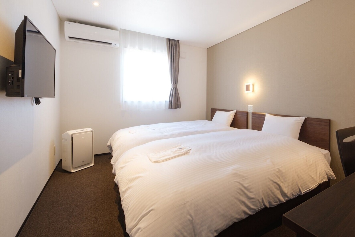 Hotellihuone, jossa on kaksi sänkyä, televisio, ilmastointilaite, ikkuna verhoilla ja yöpöytä.