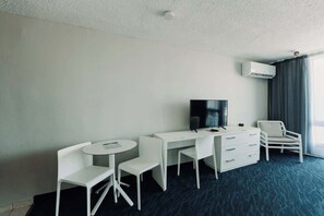 A hotel room with a desk, two chairs, a television, a dresser, and an air conditioning unit. - Oceanview Studio w/Balcony • Ocean Breezes + A/C (Carolina)