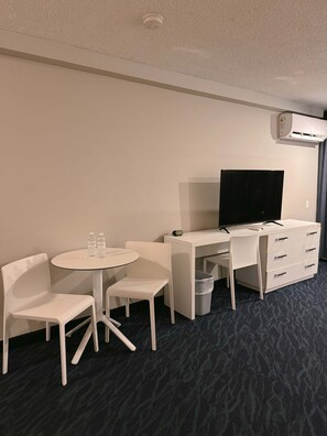 Ein Hotelzimmer mit Schreibtisch, zwei Stühlen, Fernseher, Klimaanlage und Mülleimer.